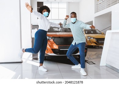 Young Afro Couple In Face Masks Dancing, Celebrating Purchase Of New Car At Auto Dealership During Covid Lockdown. Excited Black Family Buying Automobile At Showroom Store