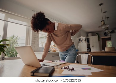 Young afro woman standing suffering from back pain while working on laptop at home