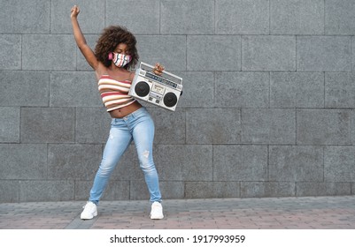 Young Afro woman wearing face mask dancing outdoor while listening to music with wireless headphones and vintage boombox stereo - Powered by Shutterstock