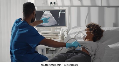 Young Afro-american Pregnant Woman In Safety At Ultrasound Office In Modern Clinic. Portrait Of Young Male Doctor Wearing Safety Mask And Gloves Using Ultrasound Machine To Check Pregnant Woman.