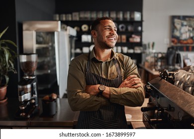 Young afro-american small coffee shop owner standing behind counter wearing apron with crossed arms looking away