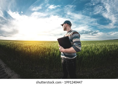 A young agronomist holds a folder in his hands on a green wheat field. A farmer makes notes on the background of agricultural land during sunset. Man in a cap with a folder of documents