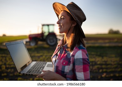 Young Agronomist Using Computer Innovation In Food Production. Female Farmer Standing In Field With Tractor Machinery In Background. 