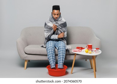 Young Ailing African American Man In Grey Plaid Scarf Sit On Sofa Soaking Feet In Warm Water Isolated On Plain Gray Background Studio Portrait. Healthy Lifestyle Ill Sick Disease Treatment Concept.