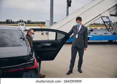 Young Airport Employee Opening The Car Door For A Man In Eyeglasses And A Protective Mask