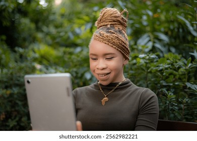 Young albino african american woman wearing a golden african continent necklace is smiling while using a tablet and sitting on a bench in a park. She's wearing a khaki turtleneck and has braided hair - Powered by Shutterstock