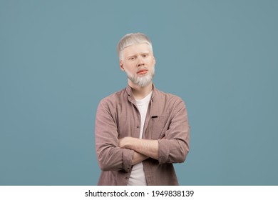 Young albino blond man posing with crossed arms, wearing casual clothes and looking at camera over turquoise studio background. Guy with serious face expression. Albinism concept
