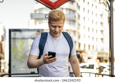 Young albino man leaving a Madrid metro stop using his smartphone and listening to music with headphones. He wears white clothes and carries a backpack. - Powered by Shutterstock