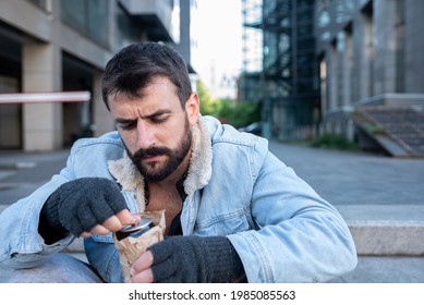 Young Alcoholic Homeless Man Sitting On The Street Opening Beer Can Hidden In The Paper Bag To Drink And Get Drunk After He Got Fired From The Job And Fell In To Depression, Social Documentary Concept