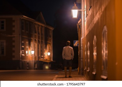 Young Alone Man Slowly Walking On Sidewalk Under Street Lights At Old Town In Dark Summer Night. Peaceful Atmosphere. Spending Time Alone. City Life. Back View. 