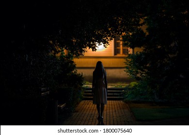 Young alone woman in dress walking on sidewalk through dark park to home in summer black night. Scary moment and gloomy atmosphere. Back view.