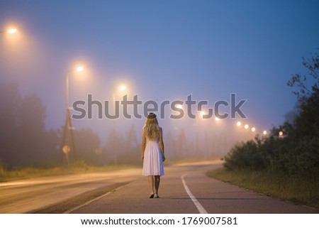 Young alone woman in white dress slowly walking on long sidewalk under street lights in dark summer night. Peaceful moment. Spending time alone. Back view.