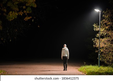 Young alone woman in white jacket standing on road under lamp light in autumn night. Back view. Road to nowhere concept. Empty place for text, quote or sayings.