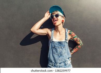 Young alternative girl wearing cap and sunglasses standing isolated on grey wall on the city street looking aside showing tongue playful - Powered by Shutterstock