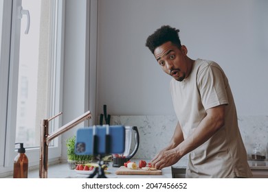 Young Amazed African American Man 20s In Casual Clothes Prepare Sweet Breakfast Cut Fruit Watch Series On Mobile Cell Phone Cooking Food In Indoor Kitchen At Home Alone Healthy Diet Lifestyle Concept