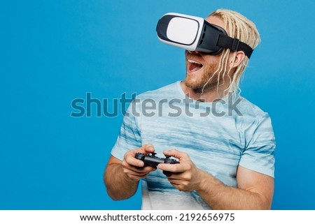Young amazed blond man with dreadlocks 20s he wear white t-shirt hold in hand play pc game with joystick console watching in vr headset pc gadget isolated on plain pastel light blue background studio
