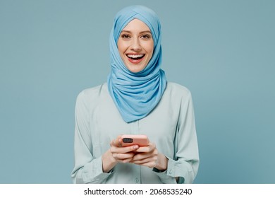 Young Amazed Happy Arabian Asian Muslim Woman In Abaya Hijab Hold In Hand Use Mobile Cell Phone Isolated On Plain Blue Background Studio Portrait. People Uae Middle Eastern Islam Religious Concept.