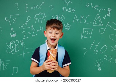 Young Amazed Male Kid School Boy 5-6 Years Old In T-shirt Backpack Biting Sandwich Eating Lunch Isolated On Green Wall Chalk Blackboard Background. Childhood Children Kids Education Lifestyle Concept.