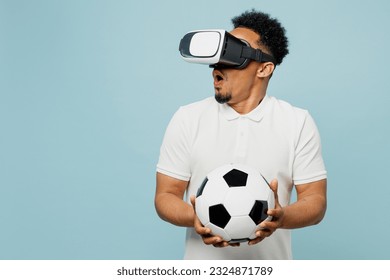 Young amazed man fan wearing basic t-shirt cheer up support football sport team hold in hand soccer ball watching in vr headset pc gadget watch tv live stream isolated on plain blue color background - Powered by Shutterstock