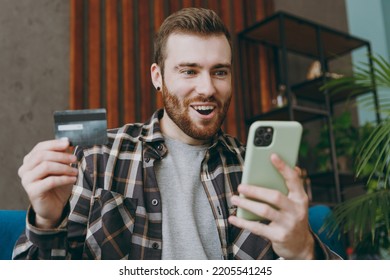 Young Amazed Man He Wear Brown Shirt Use Mobile Cell Phone Hold Credit Bank Card Shopping Online Order Delivery Booking Tour Sitting On Blue Sofa In Own Living Room Apartment Stay Home Indoor Flat On