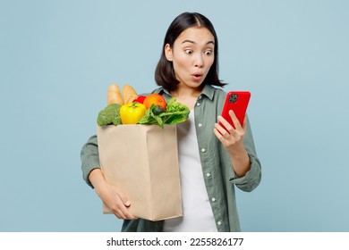 Young amazed woman wear casual clothes hold brown paper bag with food products use mobile cell phone isolated on plain blue cyan background studio portrait. Delivery service from shop or restaurant - Powered by Shutterstock