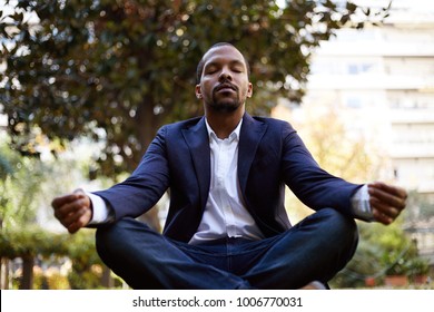 Young American African Businessman In Informal Clothes Meditating In Lotus Pose Taking A Deep Breath Outside Corporate Office.Business Yoga And Stress Free Environment.Peace Of Mind Concept.Blurred