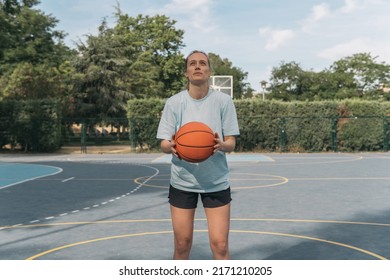 Young American Girl Basketball Player Performing A Free Throw. Sporty Blonde Woman Holding An Orange Ball And Ready To Throw It Into The Basket To Score In A Street Basketball Game On A Blue Court.