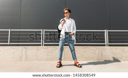 Young American man model in stylish clothes in sunglasses in red leather sandals with a vintage black bag stands on a bright sunny day near a gray building. Urban hipster guy. Fashionable menswear.