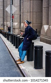 Young American Man Traveling In New York, Wearing Blue Long Overcoat, Scarf, Cuffed Knit Beanie Hat With Pom Pom, Jeans, Leather Shoes, Carrying Back Bag, Sitting On Street, Texting On Cell Phone.