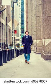 Young American Man Traveling In New York, Wearing Blue Long Woolen Overcoat, Scarf, Jeans, Cuffed Knit Hat, Carrying Back Bag, Hands In Pockets, Walking On Vintage Street With High Buildings In Winter