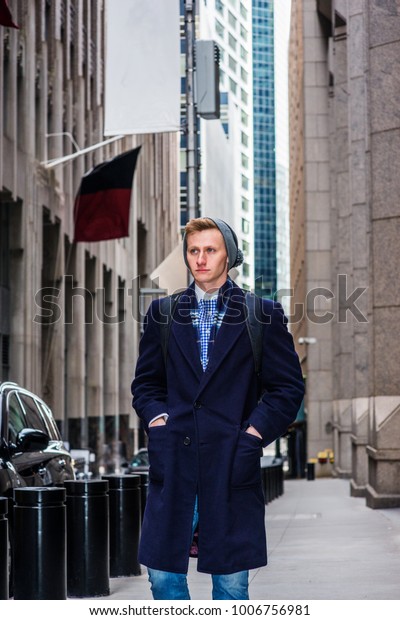 Young\
American man traveling in New York, wearing blue long overcoat,\
scarf, jeans, knit hat, carrying back bag, hands in pockets,\
walking on vintage street with high\
buildings.\
