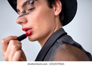 Young androgynous man with party make-up applying pomade lipstick, dressed in black shirt and hat. chic glamorous guy isolated over gray studio background. fashion, beauty, lgbtq, cosmetics - Powered by Shutterstock