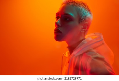 Young androgynous woman in futuristic raincoat with short hair looking away while standing under bright neon light against orange background - Powered by Shutterstock