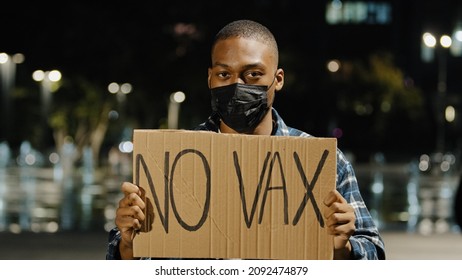 Young Angry African American Man In Black Mask Stands In City At Night Evening Holding Sign Banner With Text No VAX Guy Protest Of Covid 19 Vaccine Stop Immunization Concept. Vaccination Phobia