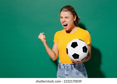 Young angry ardore woman fan wearing basic yellow t-shirt cheer up support football sport team hold in hand soccer ball watch tv live stream scream clench fist isolated on dark green background studio - Shutterstock ID 2232170039