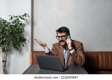 Young Angry And Frustrated Man Sitting At Cafeteria Talking On Smartphone And Argue With Home Decoration Agency After He Saw On Laptop Mail With Their Wrong Project. Unhappy CEO Of Small Business.