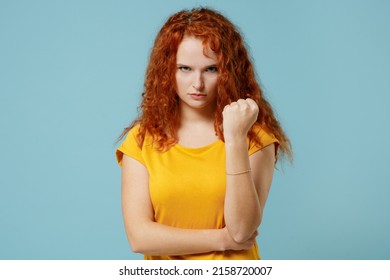 Young angry furious bad sad redhead woman 20s wearing yellow t-shirt look camera show clench fst threatening isolated on plain light pastel blue background studio portrait. People lifestyle concept