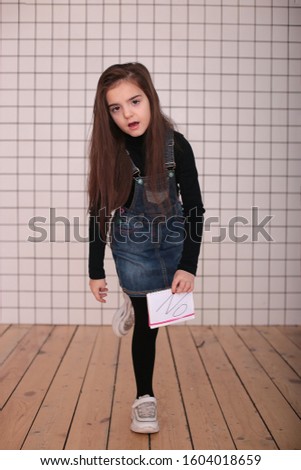 young angry playful girl of eight years with long hair in a black turtleneck and jeans sundress with the sign "NO"