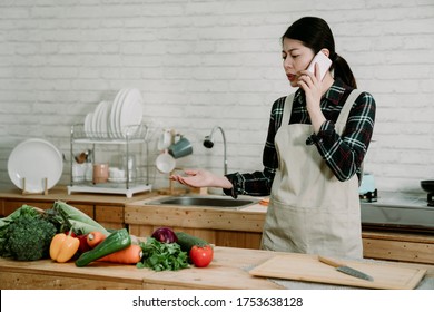 Young Angry Sad Asian Chinese Woman Housewife Talking On Cellphone Call In Bright Modern Kitchen. Many Green Vegetables On Counter Table. Mad Lady In Apron Argue Online On Mobile Phone During Cooking