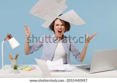 Young angry sad director employee business woman in casual shirt sit work at white office desk with pc laptop throwing up paper account documents scream shout isolated on pastel blue background studio