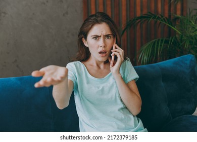 Young Angry Sad Irritated Woman She 20s Wear Casual Clothes Mint T-shirt Talk Speak On Mobile Cell Phone Conducting Pleasant Conversation Sit On Blue Sofa Indoor Rest At Home In Own Room Apartment.