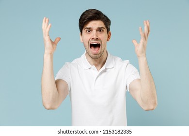 Young angry stressed indignant unshaven caucasian man 20s in white casual basic t-shirt spreading hands scream shouting look camera isolated on pastel blue background studio. People lifestyle concept