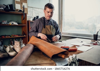 A young apprentice in a boot workshop prepares leather for further use on a large table.