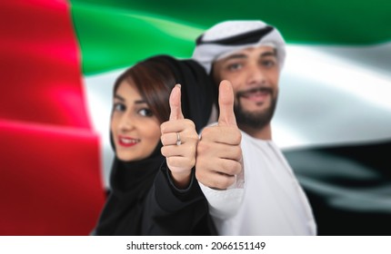 Young Arab couple showing thumbs up celebrating UAE National day, standing front UAE flag.