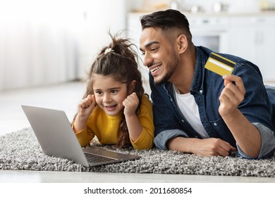 Young Arab Dad And Little Daughter With Laptop And Credit Card Making Online Shopping While Relaxing On Floor At Home, Happy Middle Eastern Family Enjoying Purchase From Internet, Closeup Shot