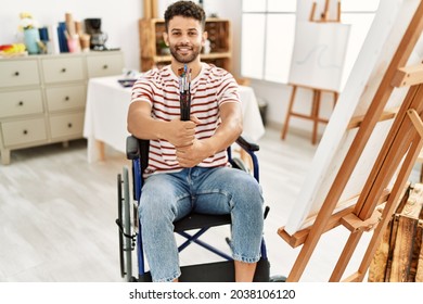 Young arab disabled artist man holding paintbrushes sitting on wheelchair at art studio.