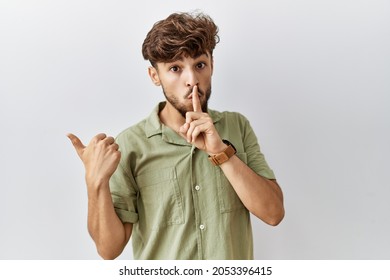 Young Arab Doctor Man Standing Over Isolated Background Asking To Be Quiet With Finger On Lips Pointing With Hand To The Side. Silence And Secret Concept. 