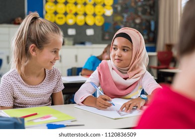Young arab girl with hijab doing exercise with her bestfriend at international school. Asian muslim school girl sitting near her classmate during lesson. Multiethnic elementary students in classroom.