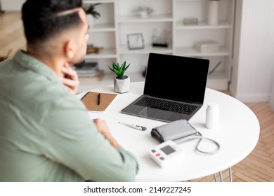Young arab guy in casual checks throat and looks at laptop with blank screen in living room interior, over shoulder view. Social distance, diagnostics, new normal and health care, covid-19 quarantine