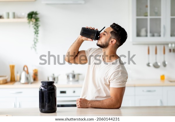 Young Arab guy drinking protein shake from bottle\
at kitchen, copy space. Millennial Eastern man using meal\
replacement for weight loss, having sports supplement for muscle\
gain. Body care concept